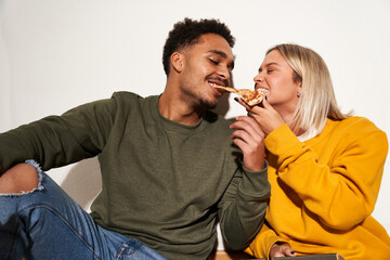 Positive multiracial couple eating pizza slice together while having fun and looking at each other