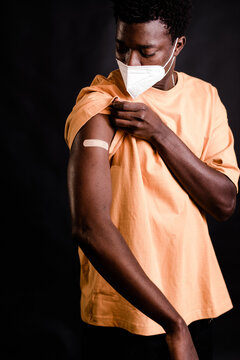 African American man with protective mask looking at adhesive bandage plaster on arm after getting the vaccination standing together on black background in a clinic during coronavirus outbreak