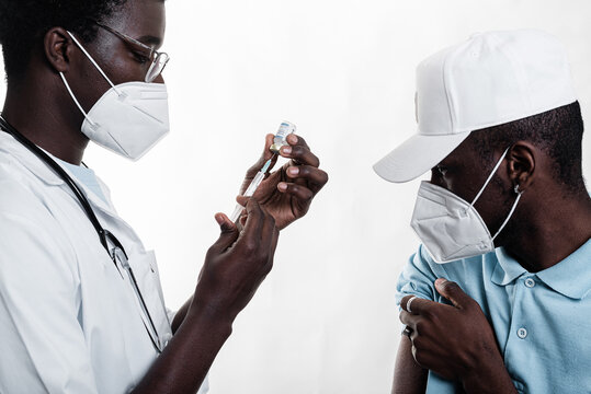 Ethnic doctor filling in syringe from bottle with vaccine preparing to vaccinate male African American patient in white background in a clinic during coronavirus outbreak
