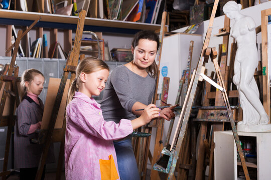 Smiling woman teacher correcting work of girl during painting class at art studio