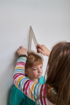Sister helping brother with measuring him height with ruler and pencil near wall