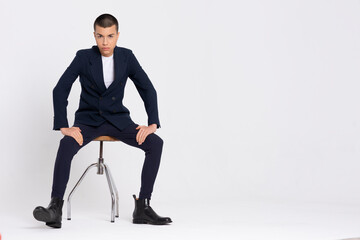Serious male model in fashionable suit sitting on white background in studio and looking at camera
