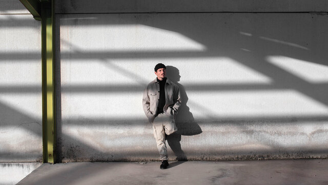 Stylish male leaning on gray shabby wall in urban area of city illuminated by sunlight and looking away