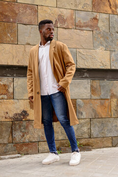 Full body of African American male wearing trendy coat standing near stone wall of building in city and looking away
