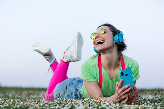 Delighted young female in colorful clothes using smartphone and listening to favorite music via headphones while lying on grassy meadow with legs crossed and looking away happily