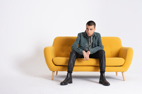 Young Handsome Male Model In Trendy Clothes Sitting On Yellow Couch On White Background In Studio And Looking Away