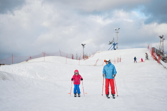 Full body faceless parent in warm sportswear and helmet teaching little kid to ski alongside snowy hill slope in winter ski resort