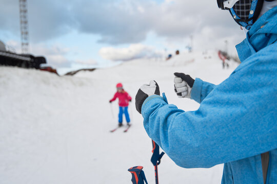Faceless parent in warm sportswear teaching little kid to ski alongside snowy hill slope in winter ski resort