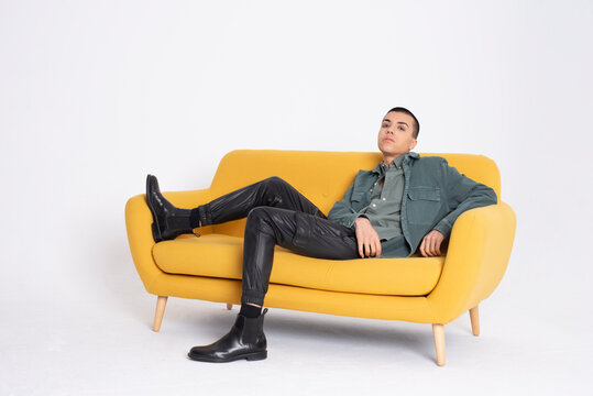 Young handsome male model in trendy clothes sitting on yellow couch on white background in studio