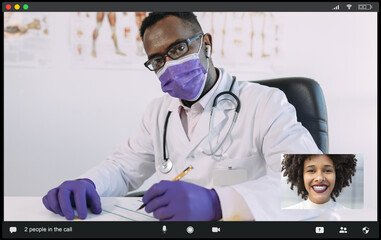 Concentrated African American male doctor in medical robe and mask looking at camera while communicating with positive female during video call