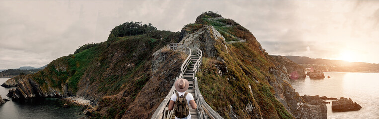 Back view of unrecognizable male tourist with backpack and hat standing on wooden pathway in a cliff and admiring scenery of mountain views in seashore during summer vacation