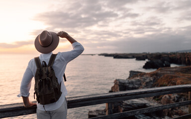 Man with backpack and hat standing on a walkway looking at the sea
