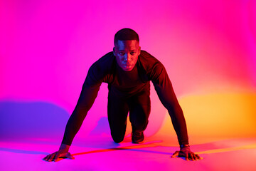 Full length concentrated fit African American male runner standing in crouch start position and looking at camera on colorful background in studio