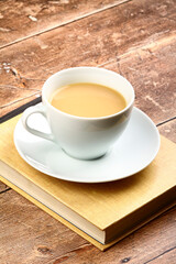 White cup and saucer filled with tea sat on a reading book on a rustic wooden table top