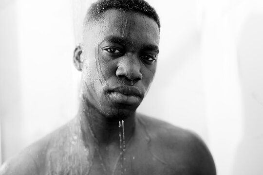 Black and white of emotionless young black guy taking shower in light bathroom and looking at camera and water on face