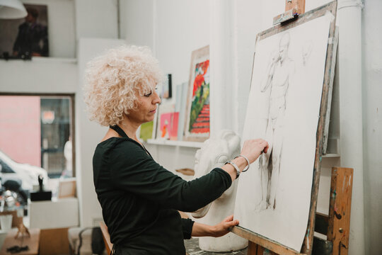 Female artist creating drawing of human with pencil while standing at easel in studio
