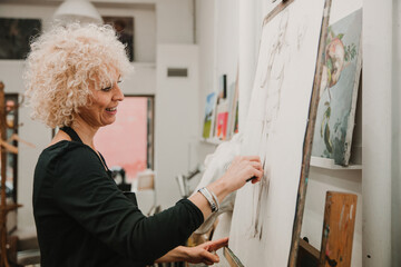 Female artist creating drawing of human with pencil while standing at easel in studio