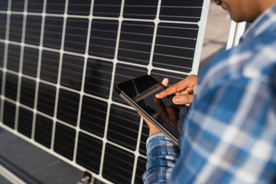 Cropped unrecognizable ethnic male technician in checkered shirt browsing tablet while standing near photovoltaic panel located in modern solar power farm