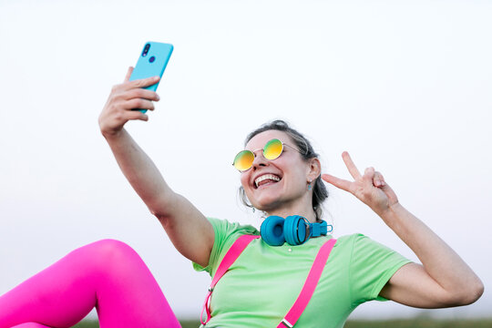 Side view joyful young female wearing bright outfit lying on lush grass taking a self portrait while making the victory symbol in countryside