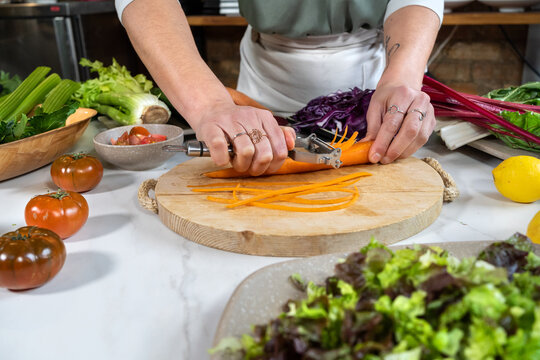 Crop unrecognizable female cutting raw carrot with peeler while preparing vegetarian food in house