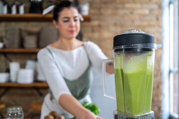 Female blending vegetables and vegetarian milk in kitchen appliance while preparing healthy green drink in house