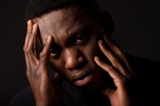 Crop unhappy young black male in black jumper with hands on face looking at camera on black background in bright studio