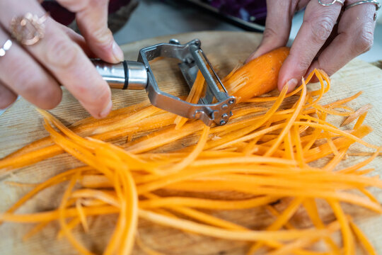 Crop unrecognizable female cutting raw carrot with peeler while preparing vegetarian food in house