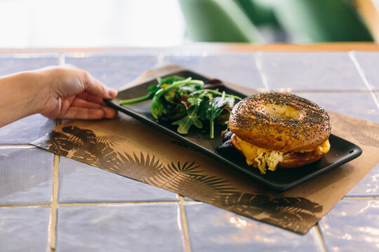 High angle of crop anonymous person serving delicious bagel sandwich with cheese and chicken on plate on table in cafe