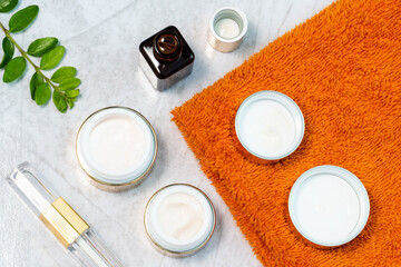 cosmetically flat lay: jars of skin care cosmetics in gold colour, an orange towel and a sprig of luscious greenery