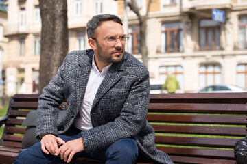 man sitting in the park on a brown bench. on the street spring, green grass, the sun is shining, good weather. on the city background. young man in glasses in a white shirt and gray coat