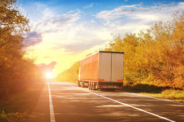 A truck with a trailer and other cars on a countryside road with trees against a sky with a sunset - 433698523