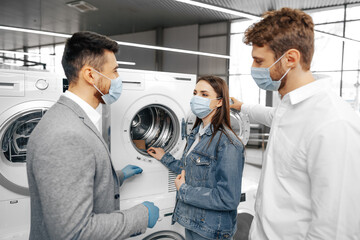 Fototapeta premium Salesman in hypermarket wearing medical mask demonstrates his clients a new washing machine