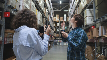 Fototapeta na wymiar Videographer woman makes clip content with concentrated young brunette guy with clipboard standing on high shelves in spacious warehouse. Close up