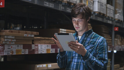 Close up. Concentrated young brunette guy employee with tablet checking goods in cardboard packages...