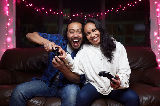 Excited ethnic couple in casual wear with joy pads playing video game together while sitting on leather couch at home