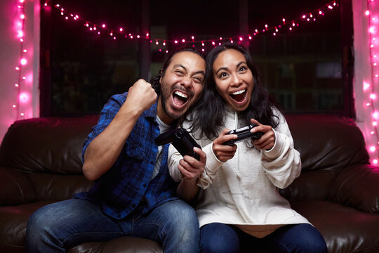 Excited ethnic couple in casual wear with joy pads playing video game together while sitting on leather couch at home