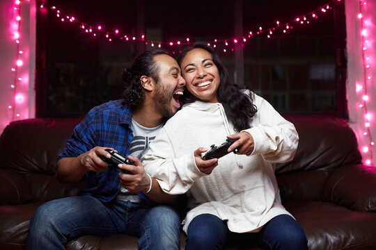 Excited ethnic couple in casual wear with joy pads playing video game together while sitting on leather couch at home