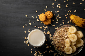 Desayuno saludable de avena con plátano, un vaso de leche y unas galletas sobre la mesa de color negro.