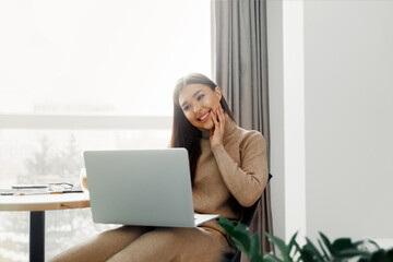 Beautiful young woman in casual clothing using laptop and smiling while working indoors. Home office during Coronavirus or Covid-19 quarantine. Communicates on internet.