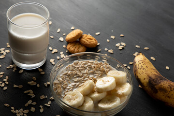 Desayuno saludable de avena con plátano, un vaso de leche y unas galletas sobre la mesa de color negro.