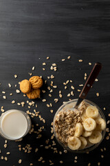 Desayuno saludable de avena con plátano, un vaso de leche y unas galletas sobre la mesa de color negro.