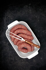 Sausages For Frying or BBQ Grilling In White Ceramic Dish And Grill Tool On Shabby Black Background, Top View. Raw Lamb Or Beef Sausages In Natural Casing In White Tray, Overhead View.