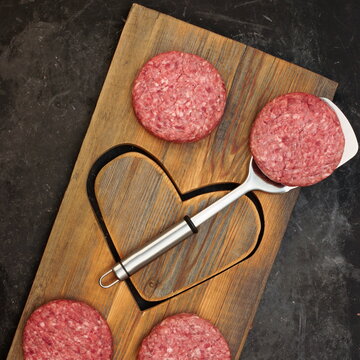 Patty of Minced Meat On Wooden Board with Shape of Heart. Raw Beef Steak Burgers on Wood Board with Heart Shape. Raw Steak Burgers Cutlets And BBQ Grill Tools Overhead View. Ground Beef Burgers.