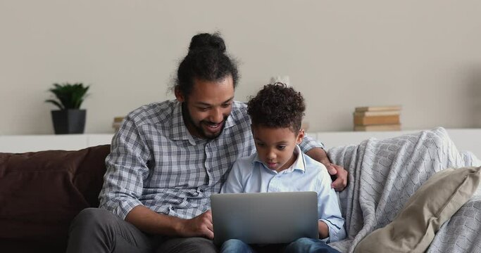 Young African dad and little son sit on sofa using wireless pc, kid and parent buying online, choose video games, learn new app, spend free time on weekend using internet and modern device concept