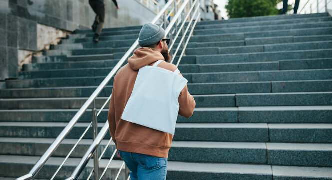 Street style portrait of young hipster man wearing brown blank hoodie with white blank textile eco shopping bag or tote bag. Mock up for design