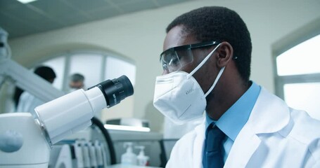 Close up of African American young male medical scientist working in laboratory on coronavirus cure. Man doctor in mask looking at blood cells analysis through microscope, experiment concept - Powered by Adobe