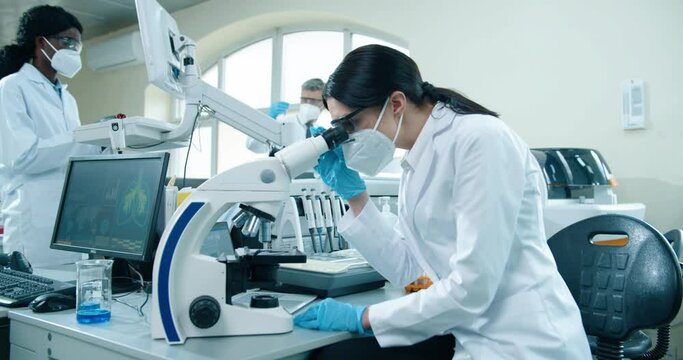 Side view of young Caucasian female medical specialist in mask and gloves sitting at laboratory using modern microscope looking analysis results. Medicine, medical researcher at lab