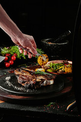 T-Bone steak serving with grilled vegetables on a black round plate on a background of greens, spices and vegetables, on a copper sheet.
