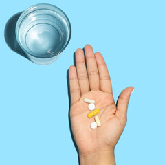 Hand holding white and yellow pills next to a glass of water flat lay in blue background