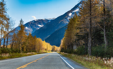 road trip on an open road in Alaska highway passing high snowcapped mountain peaks and golden yellow autumn foliage.